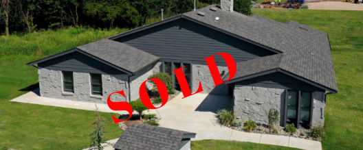 An aerial view of a large house with a gray roof surrounded by grass and trees.