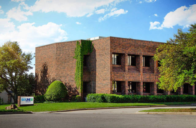Brick office building with ivy, trees, and a parking lot under a partly cloudy sky