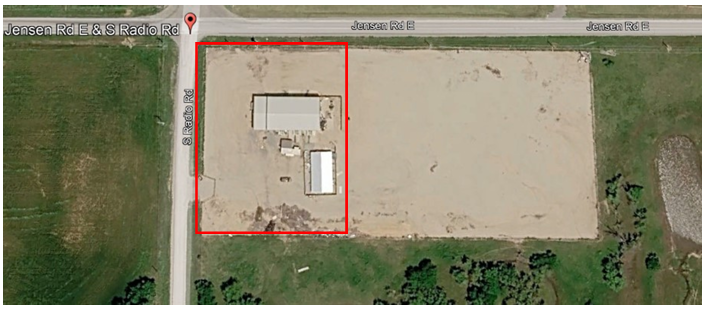 Aerial view of a rural lot outlined in red beside a road and open field, with a small building near the top left.