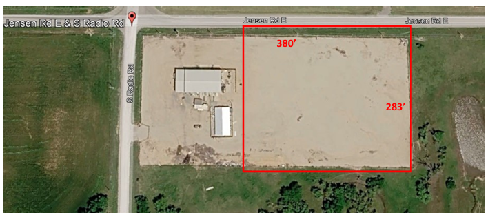 Aerial view of a vacant dirt lot outlined in red beside a road and a small building.