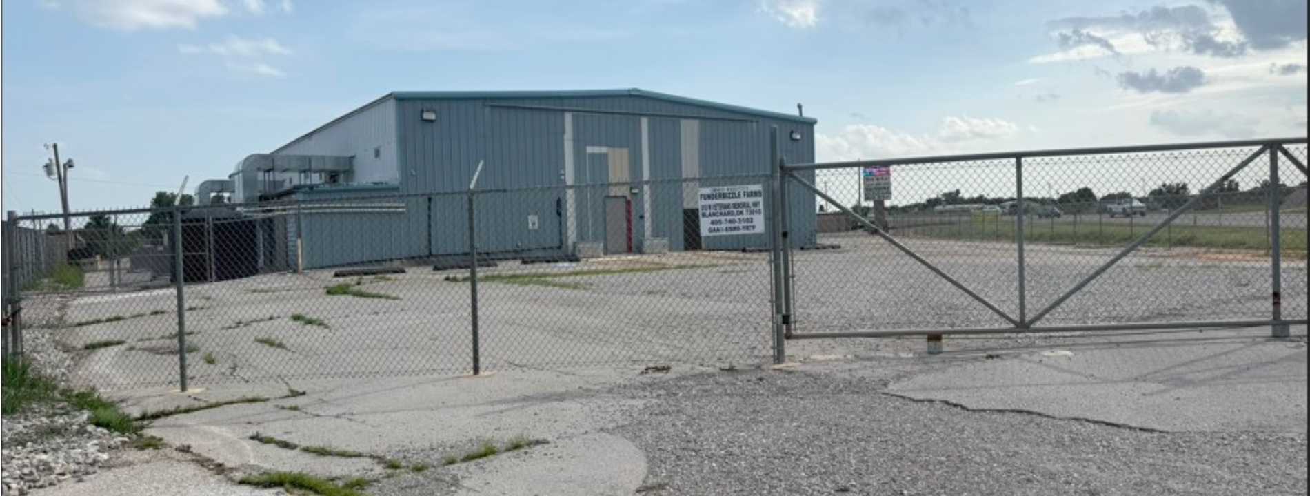 A fenced industrial building with a gate. The building is grey and the lot is gravel.