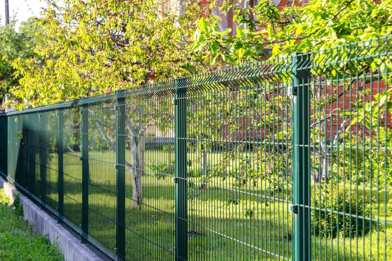 Green metal fence along a grassy yard with trees and a brick building in the background
