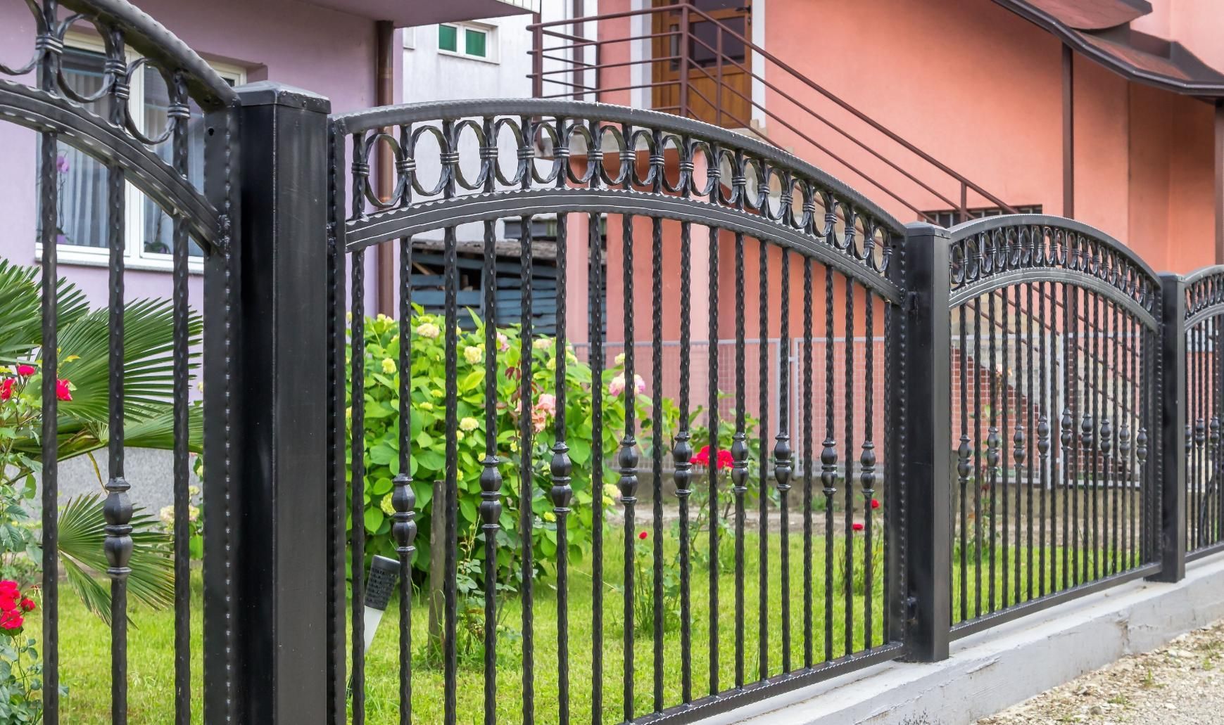 Black wrought-iron fence in front of a pink house with green shrubs and flowers