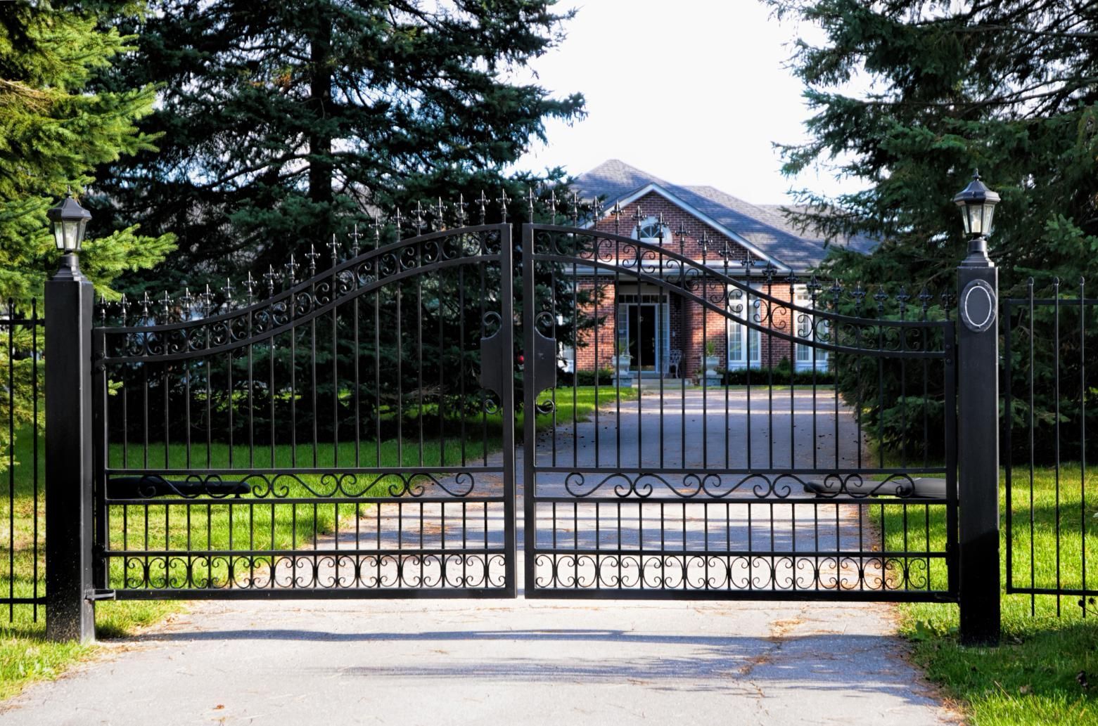 Closed black wrought-iron gate before a brick house and driveway, framed by tall trees
