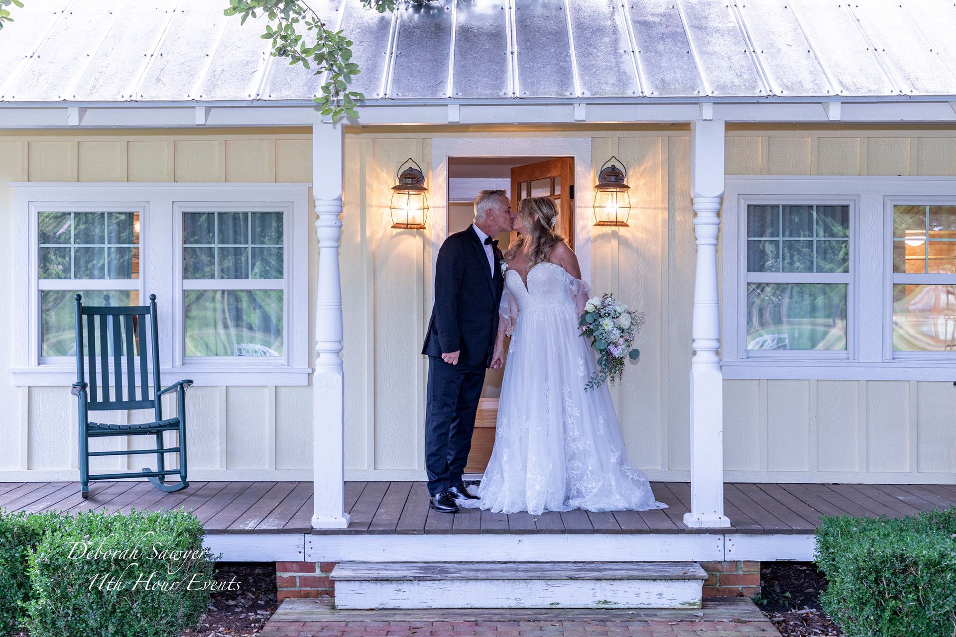 Bride and groom kissing on a porch in front of a yellow house. The groom wears a black suit, the bride a white wedding dress, and they hold a bouquet.