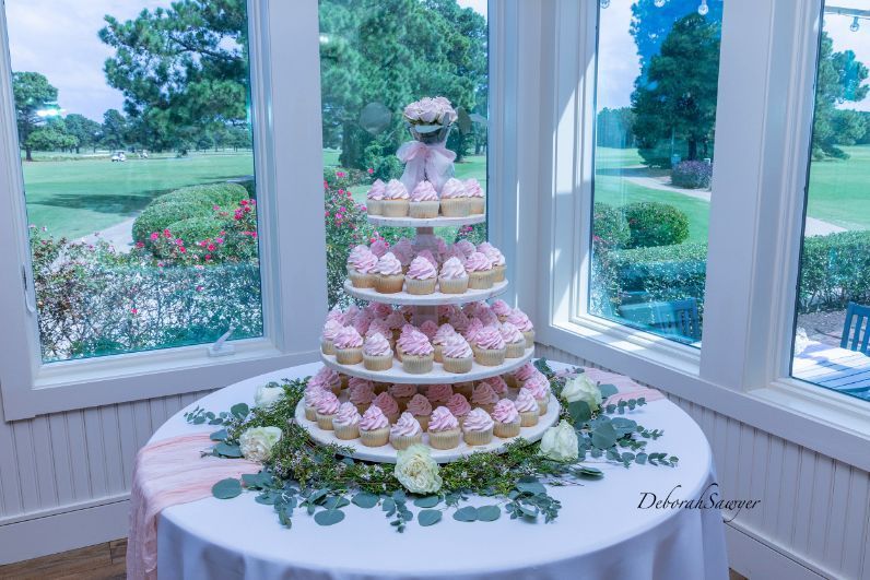 A cupcake tower with pink frosting sits on a round table decorated with greenery and flowers near a window with a golf course view.