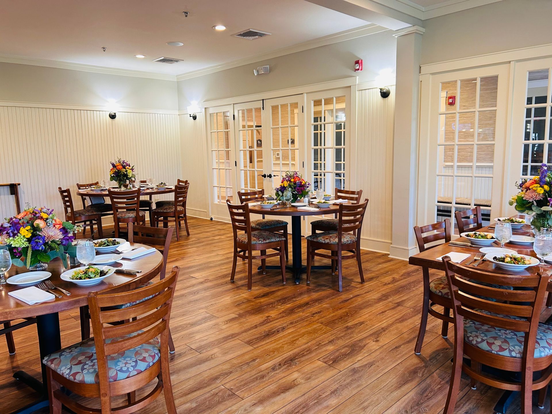 Bright dining room with round wooden tables set for meals. Floral centerpieces and chairs surround each table, with windows and doors along the walls.