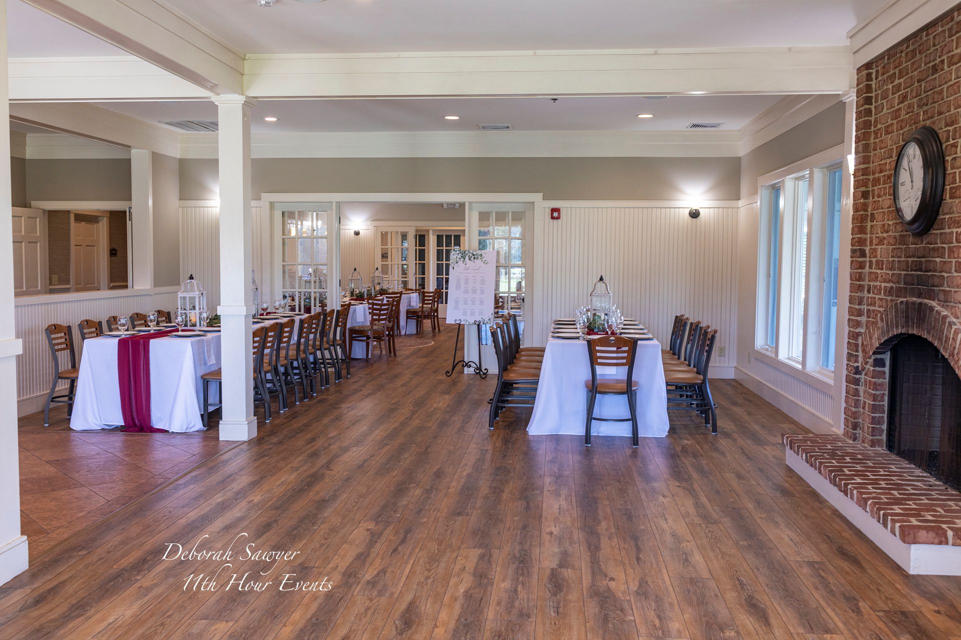 An event space with wooden floors and tables set for a celebration, featuring a brick fireplace and natural light.