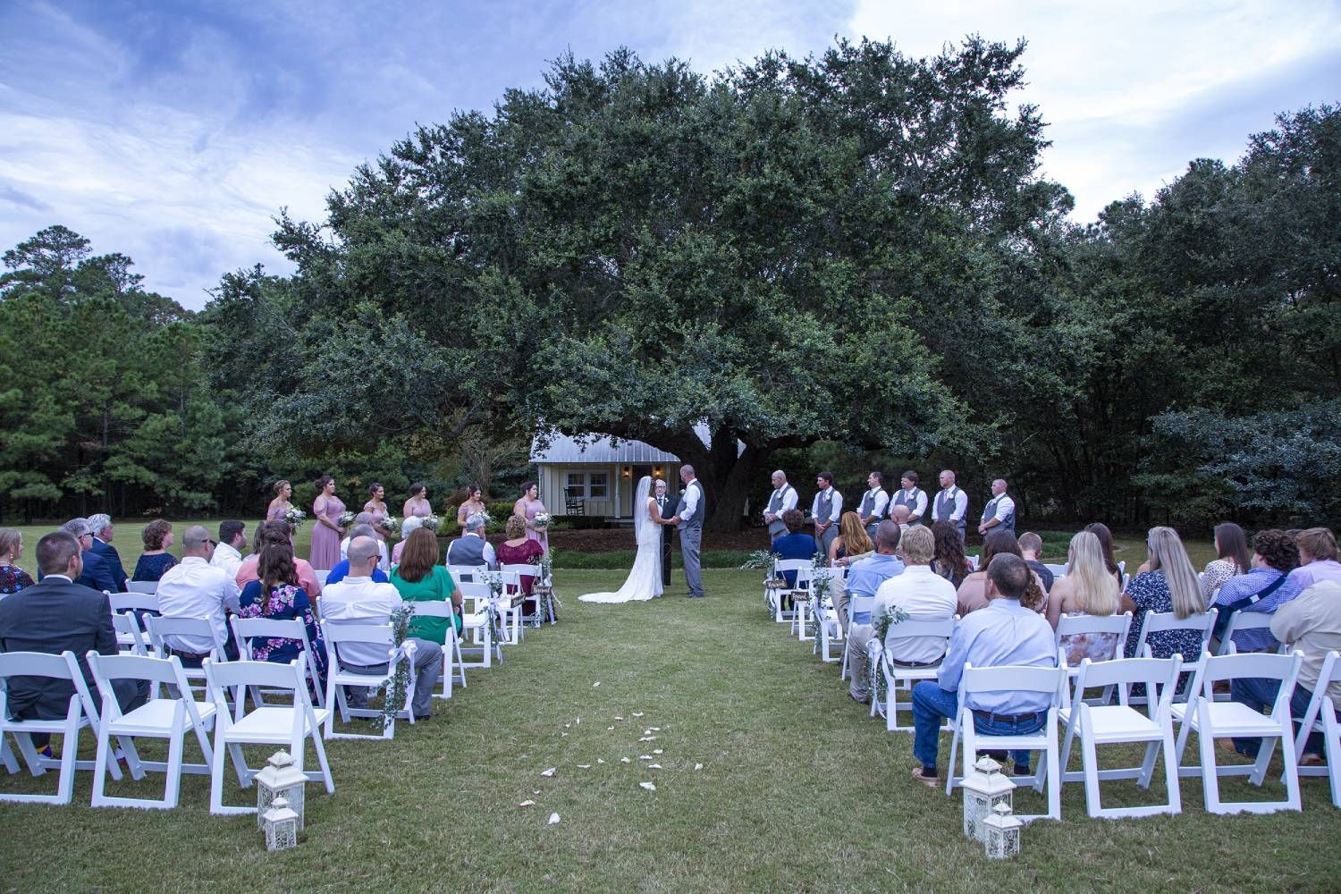Wedding ceremony outdoors: Bride and groom stand under a large tree, surrounded by guests seated in white chairs on a grassy lawn.