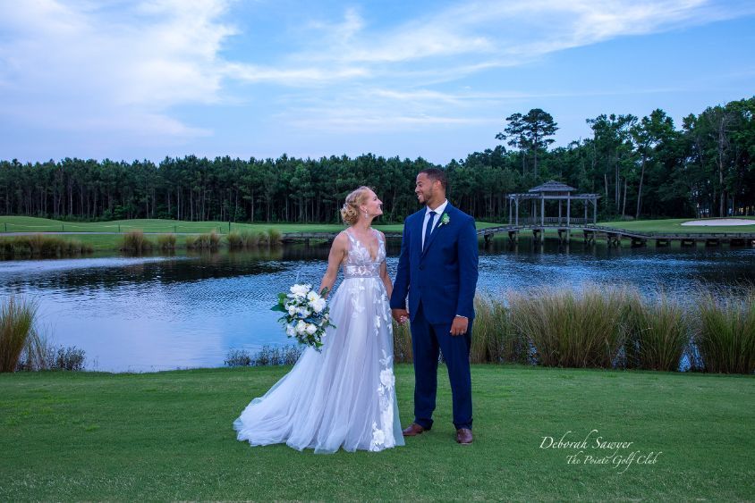 Bride and groom holding hands, smiling, and looking at each other. They stand on a golf course near a lake.
