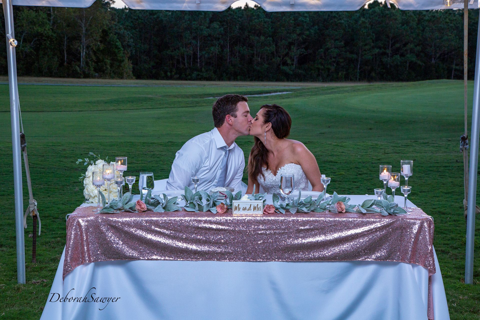 Newlyweds kissing at a reception table decorated with flowers and candles, outdoors at dusk. The table has a rose gold sequin skirt.