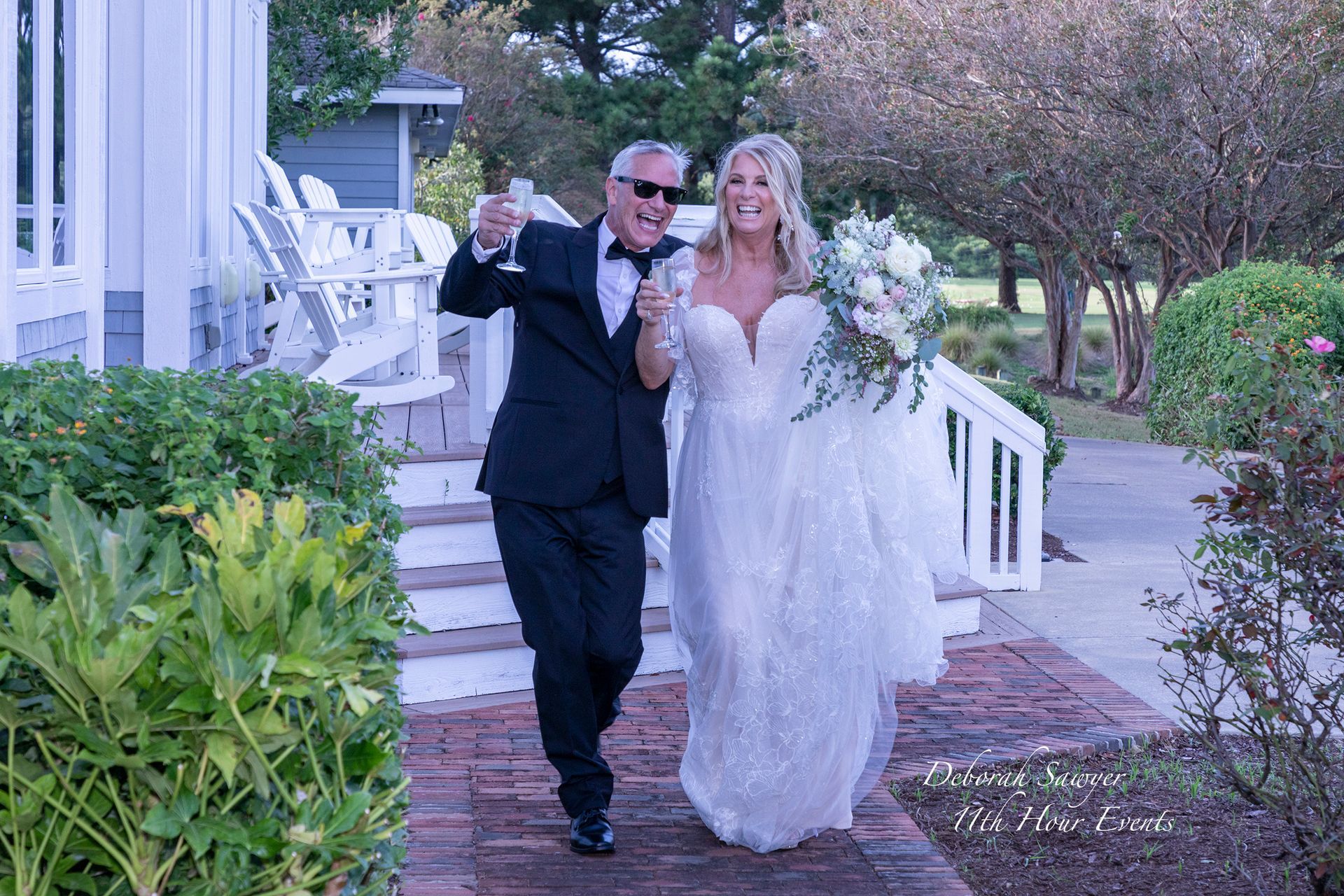 Newlyweds, a man in a tuxedo with champagne and a woman in a white wedding dress holding flowers, walking happily. Outdoors.