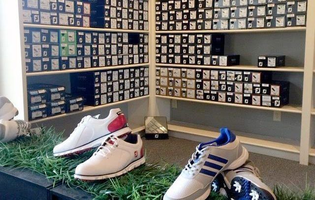 Golf shoes on display in a store, with rows of shoe boxes on shelves in the background.