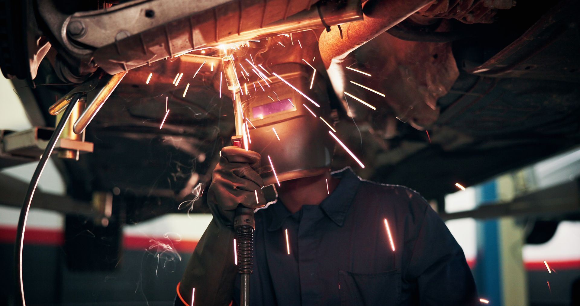Mechanic welding under a vehicle, bright sparks flying in a dim garage