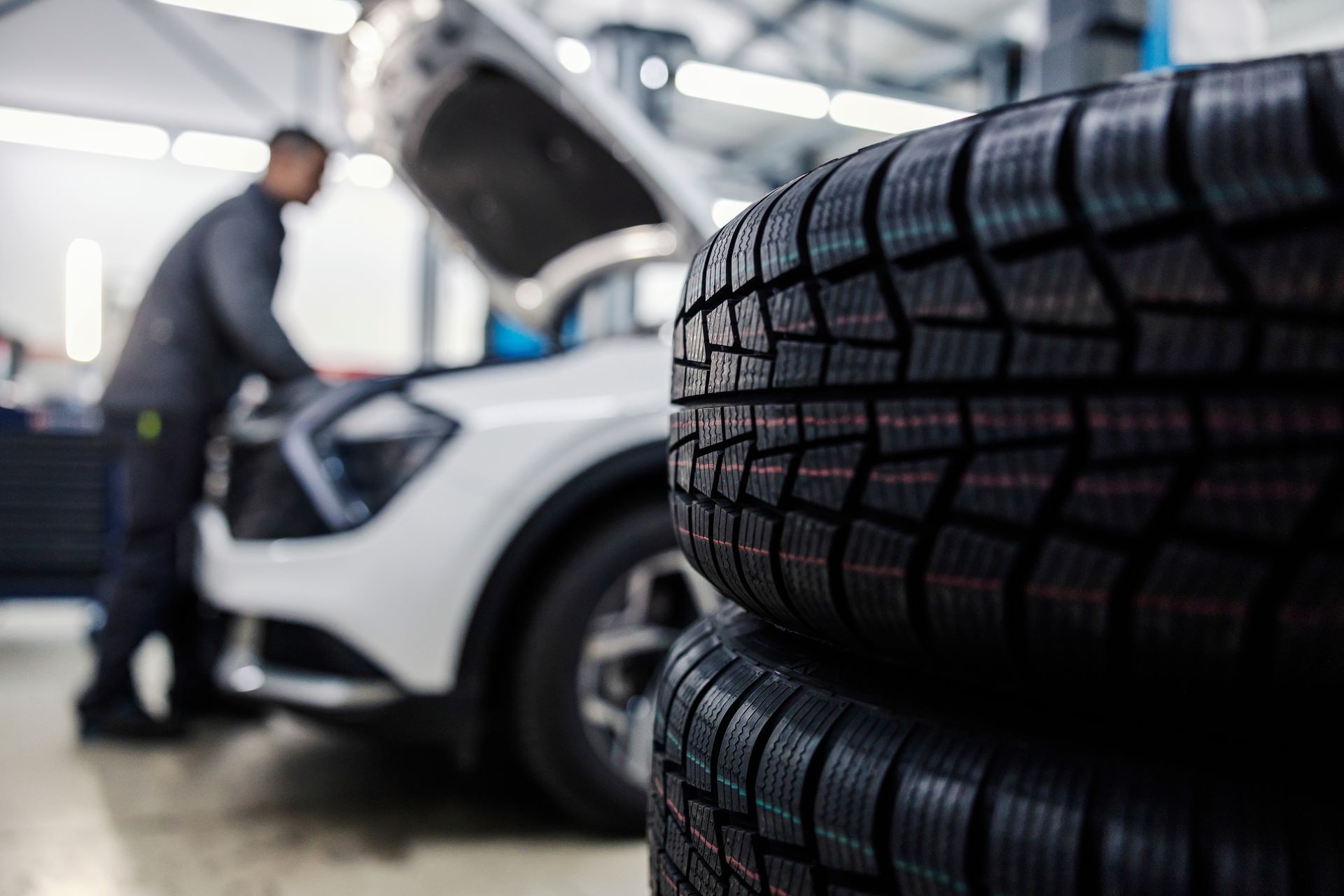 Stacked tires in foreground with a mechanic working on a white car in an auto shop
