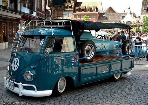 Teal Volkswagen bus hauling a classic car on a flatbed truck in a cobblestone street