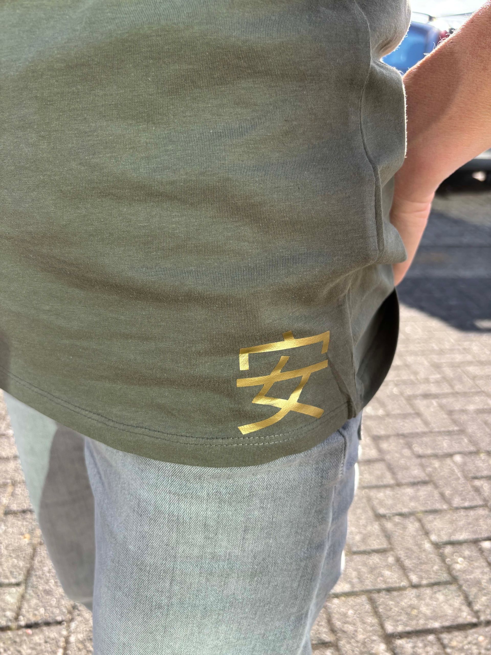Een persoon staat buiten in een groen T-shirt met een gouden vogelprint en een spijkerbroek, met de handen in de zakken.