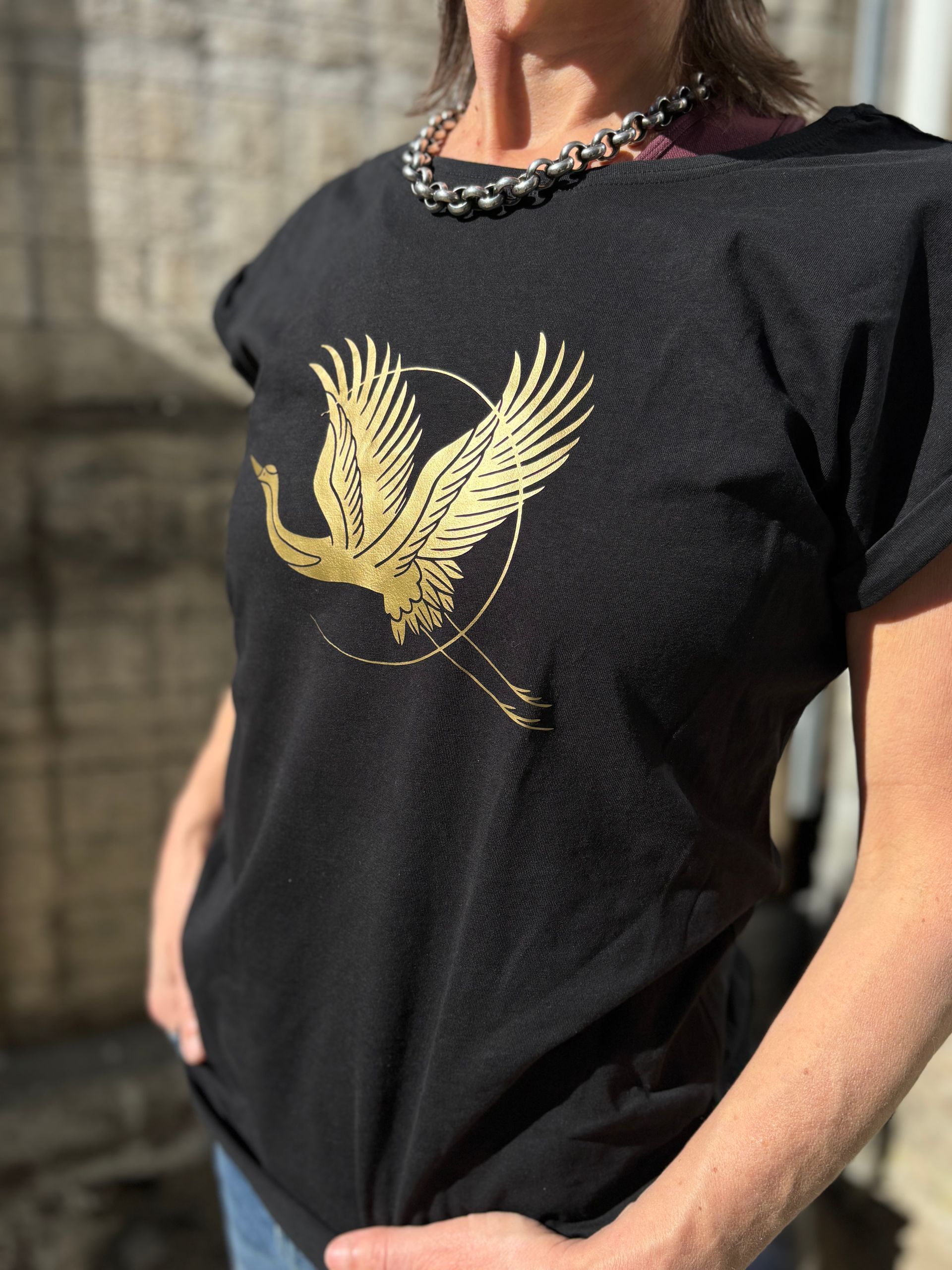 Een persoon draagt ​​een zwart T-shirt met een gouden vogelmotief en een kralenketting, buiten.