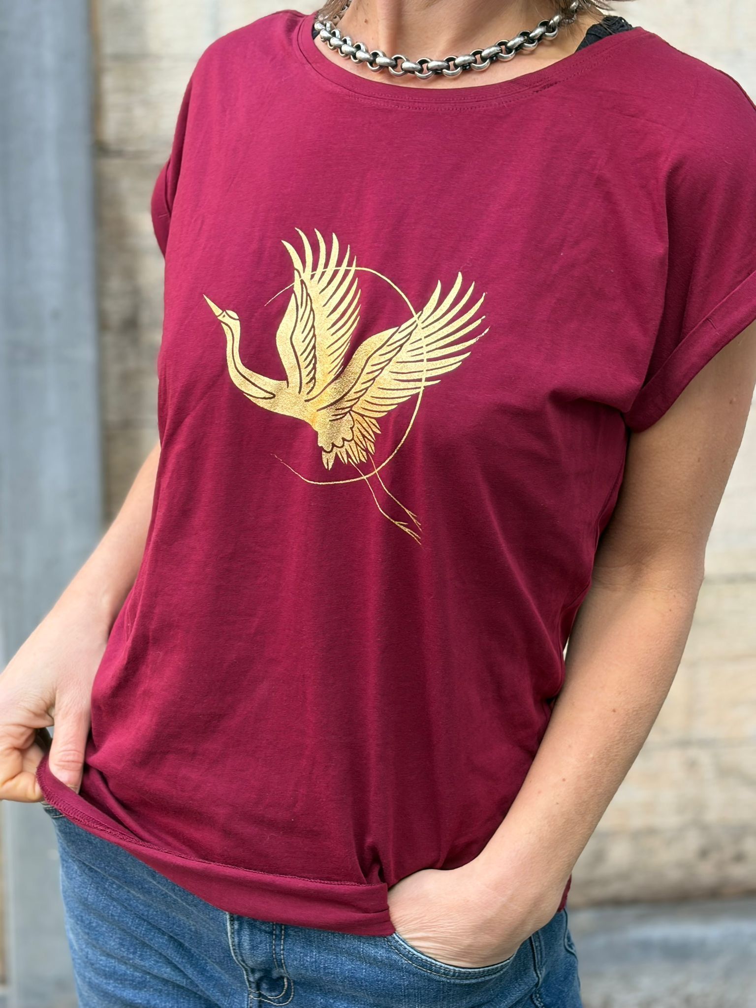 Persoon met een magenta T-shirt met een gouden vogelprint en een blauwe spijkerbroek aan, handen in de zakken.