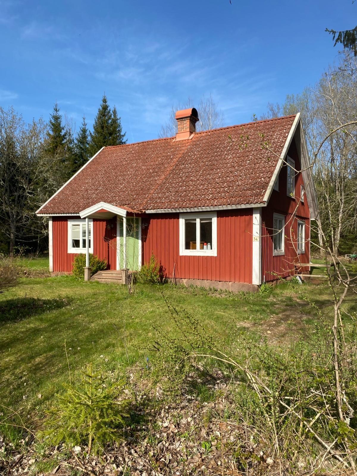 Rood houten huisje met witte kozijnen en een steil pannendak in een grasrijke tuin onder een blauwe hemel.