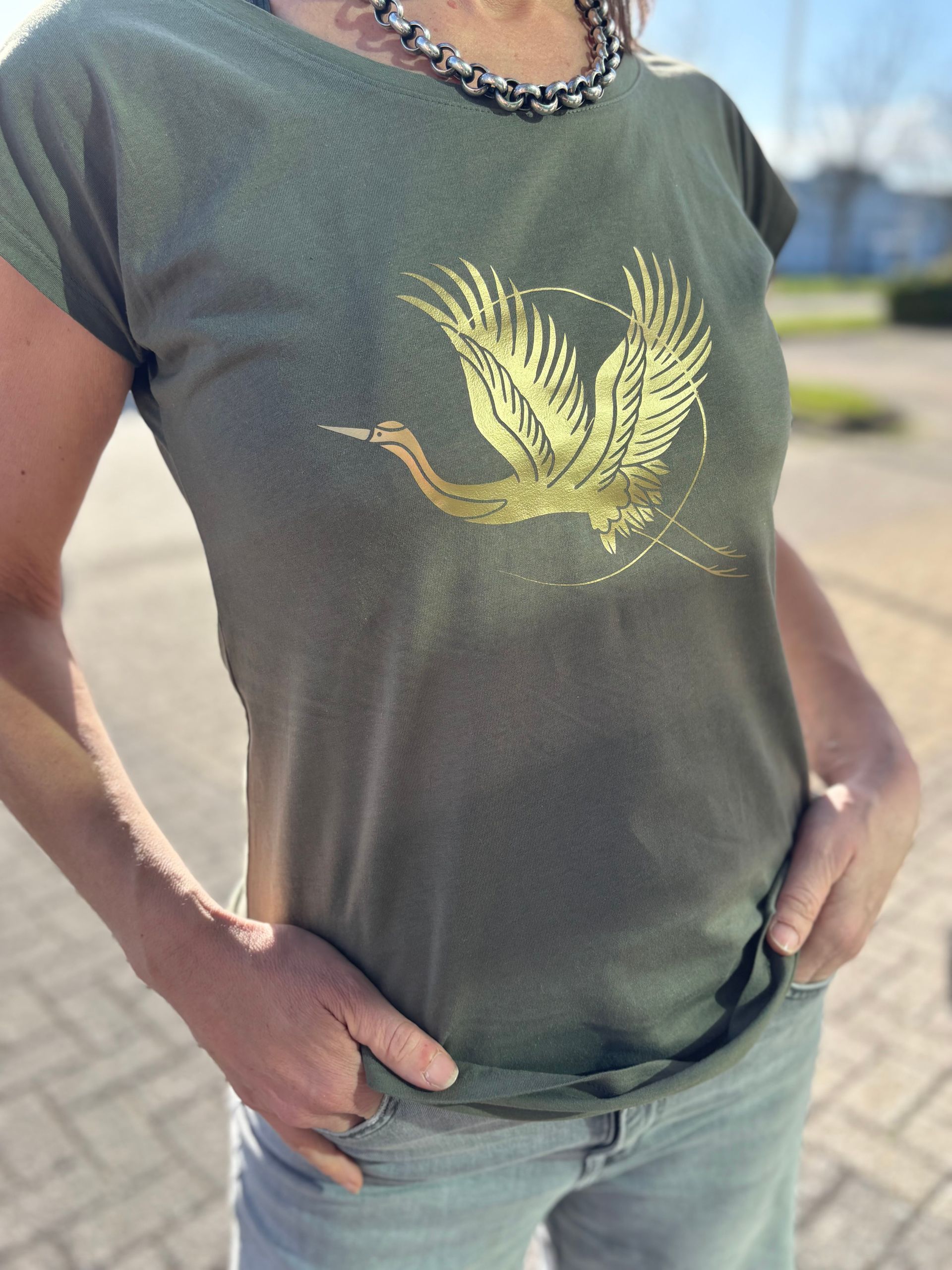 Een persoon staat buiten in een groen T-shirt met een gouden vogelprint en een spijkerbroek, met de handen in de zakken.