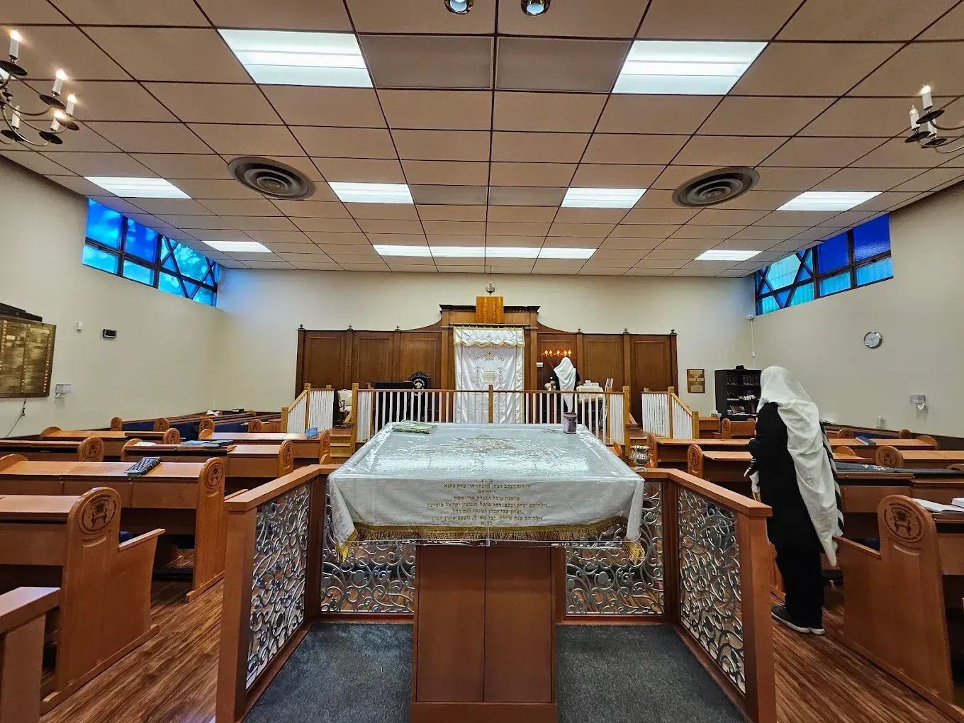 Inside a synagogue, a white-draped table stands before the ark, with wooden pews and stained-glass windows.