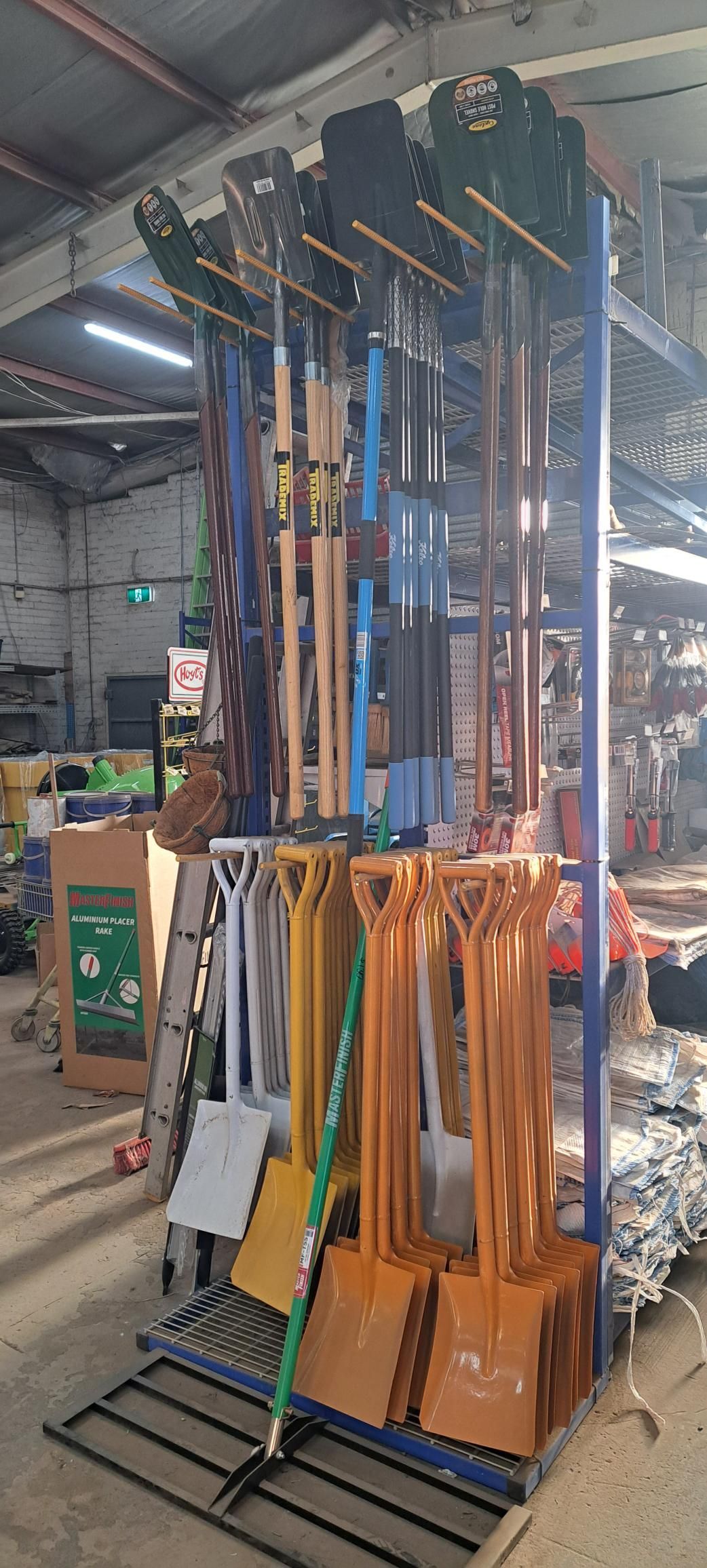 A tall metal rack holding gardening tools, including shovels, rakes, and hoes, inside a warehouse.