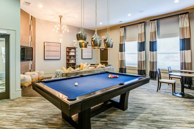 A blue billiards table sits in a modern game room with pendant lights, a sofa, and large windows with striped curtains.