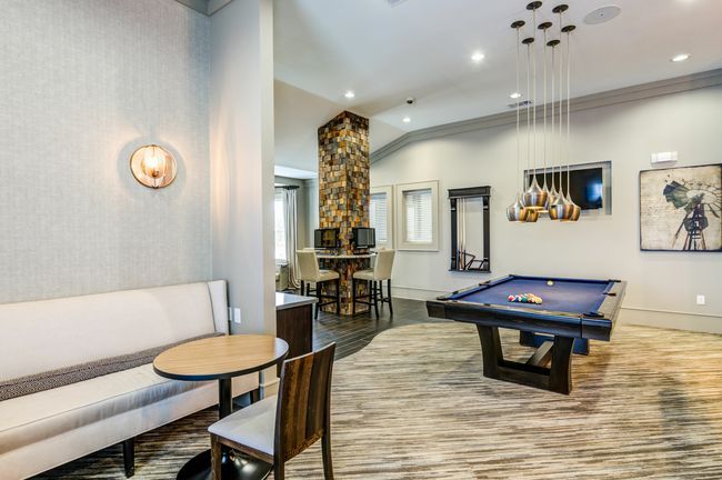 A game room featuring a billiards table, a small bistro table with chairs, and a central column with computer stations.