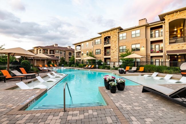 A bright outdoor pool at a modern apartment complex with tan stone buildings, lounge chairs, and umbrellas at sunset.Reli Log