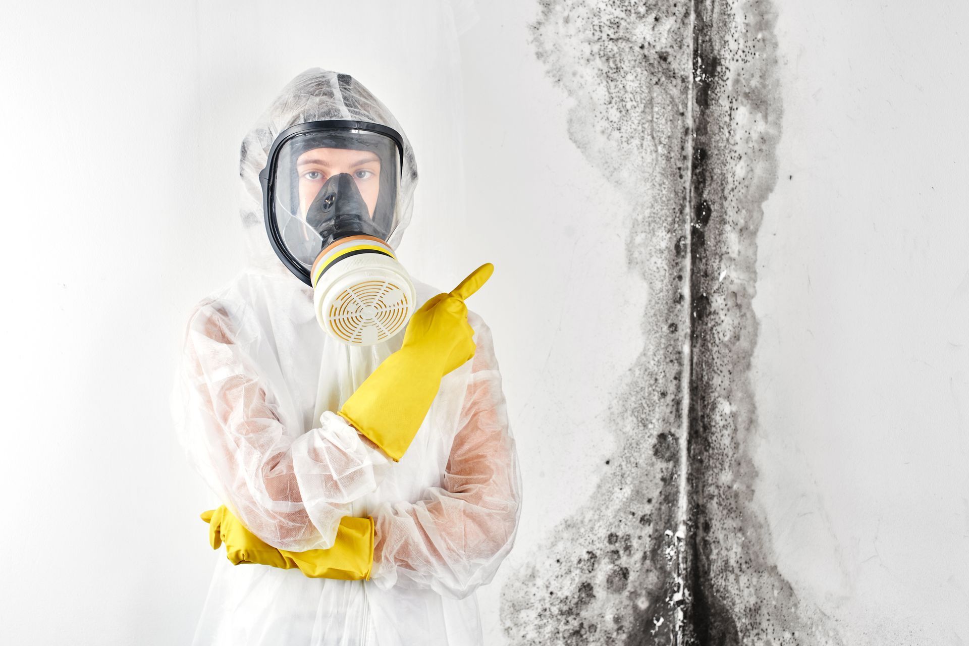 A person wearing a gas mask and yellow gloves is standing in front of a wall covered in mold.