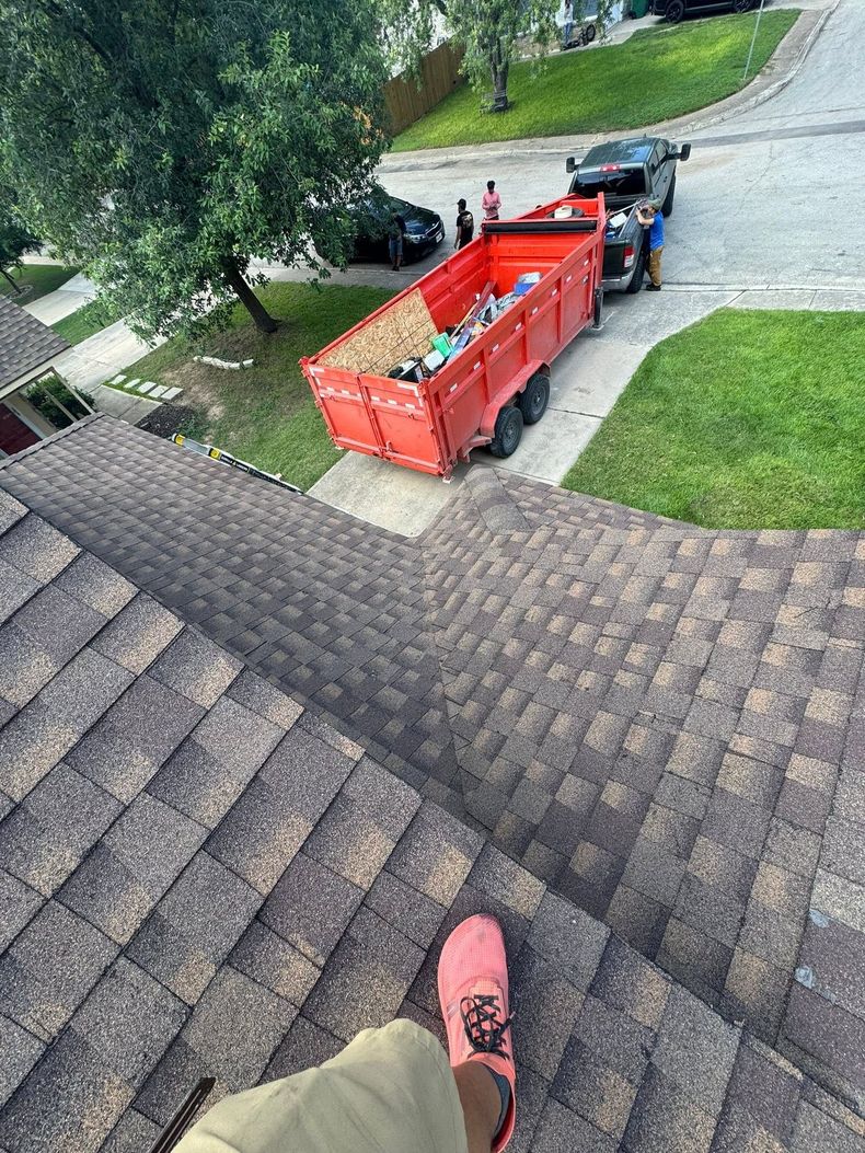 View from a shingled roof toward a red dumpster on a driveway, with one shoe visible at the bottom edge