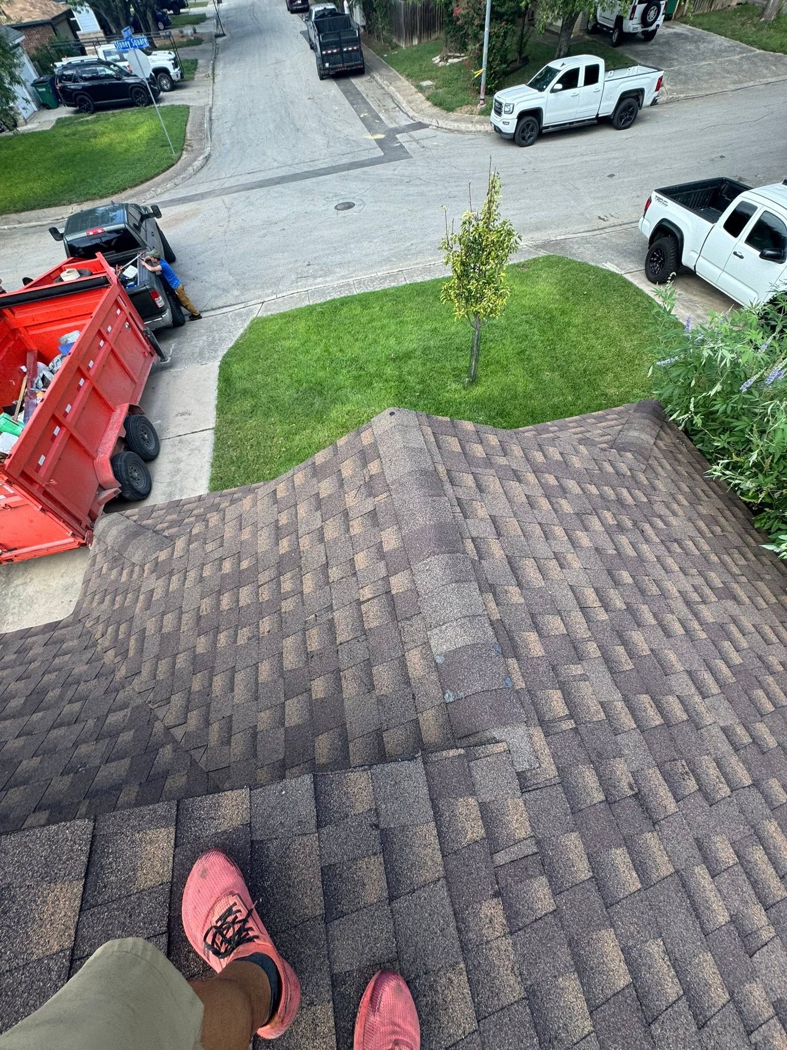 View from a shingled roof onto a driveway with parked trucks, a red dump trailer, and a small tree in the yard
