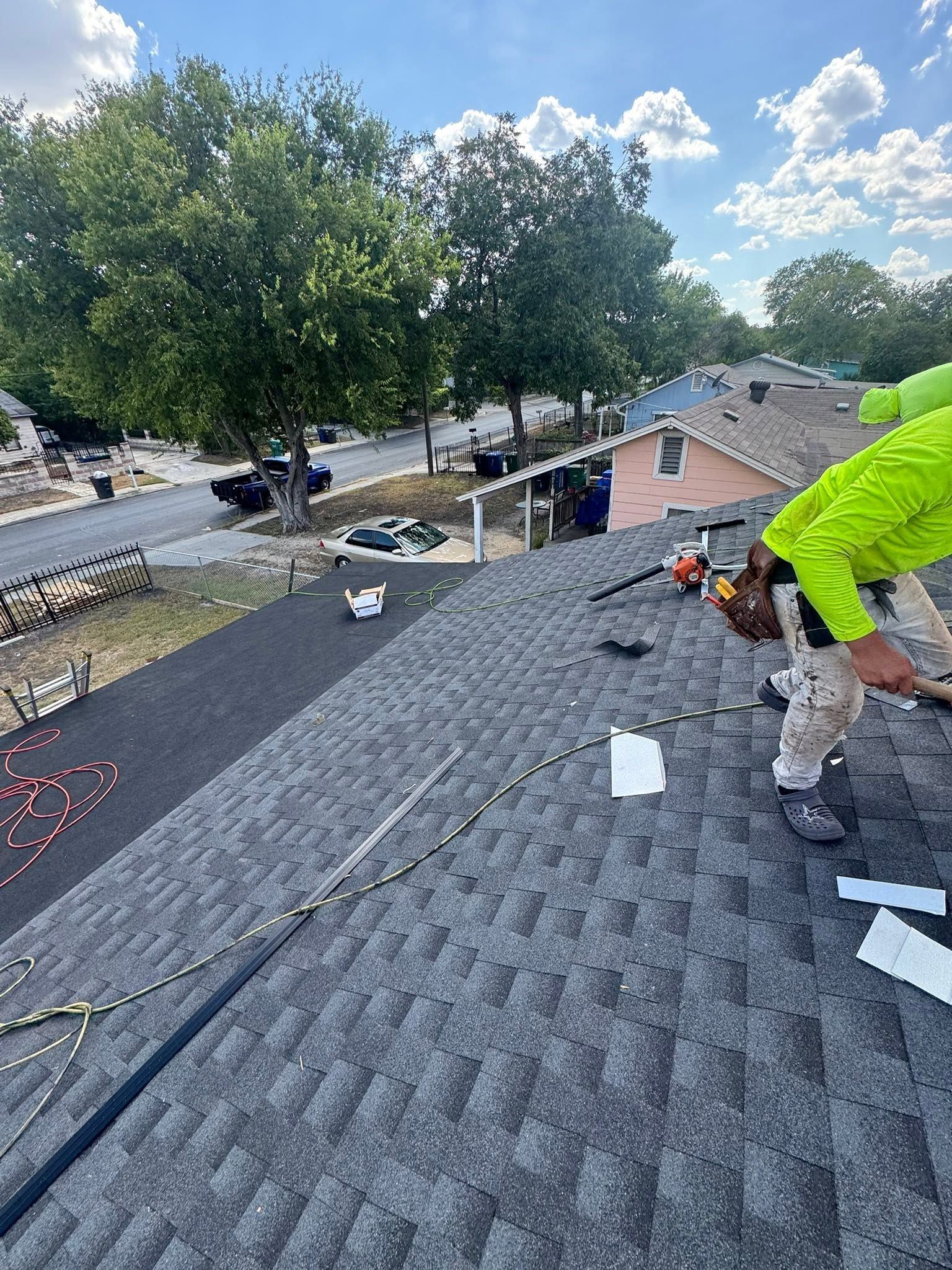 Worker on a shingled roof near trees, installing roof materials under a blue sky.