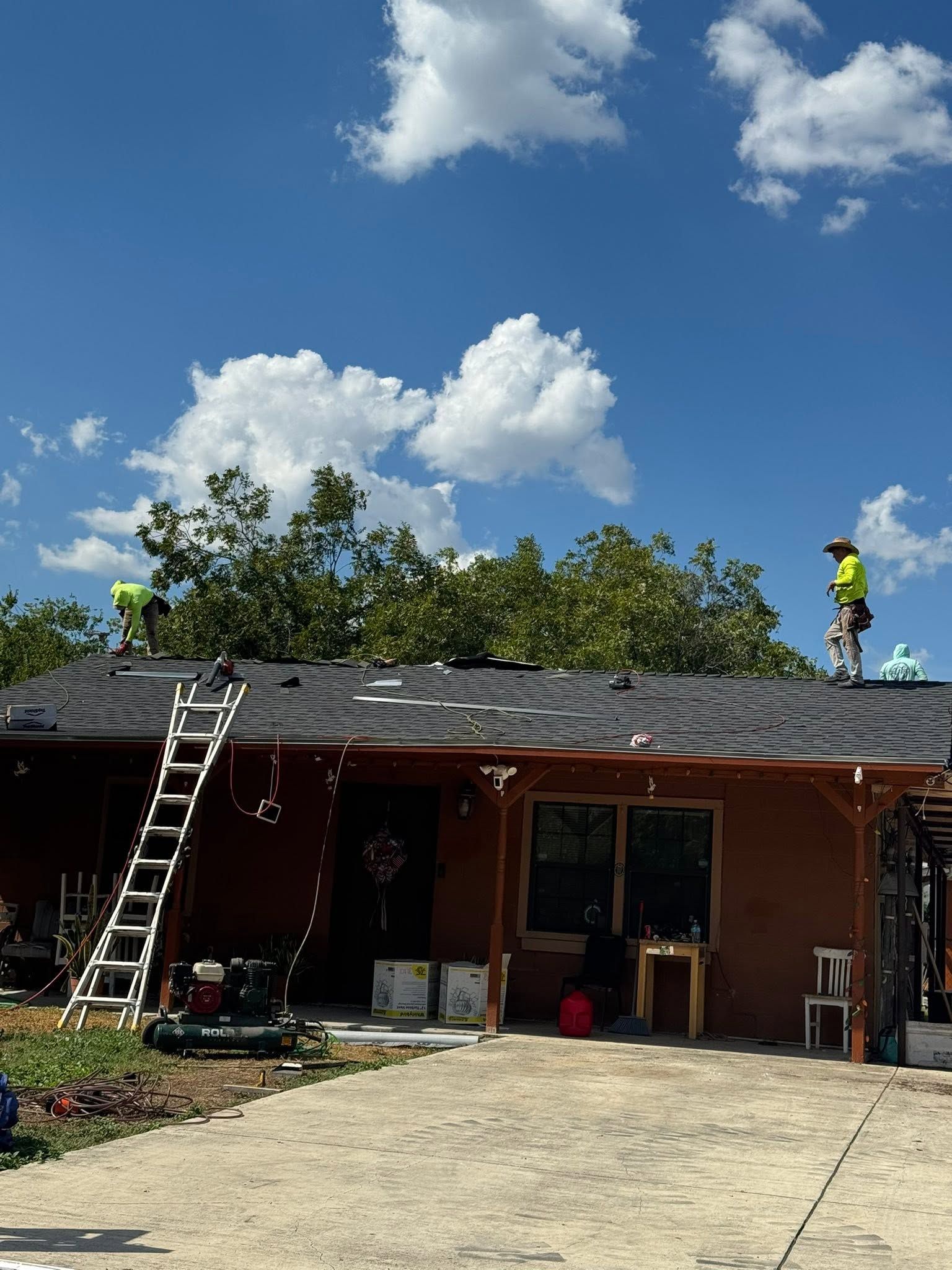 Worker on a roof repairing shingles on a house, with a ladder and clear blue sky.