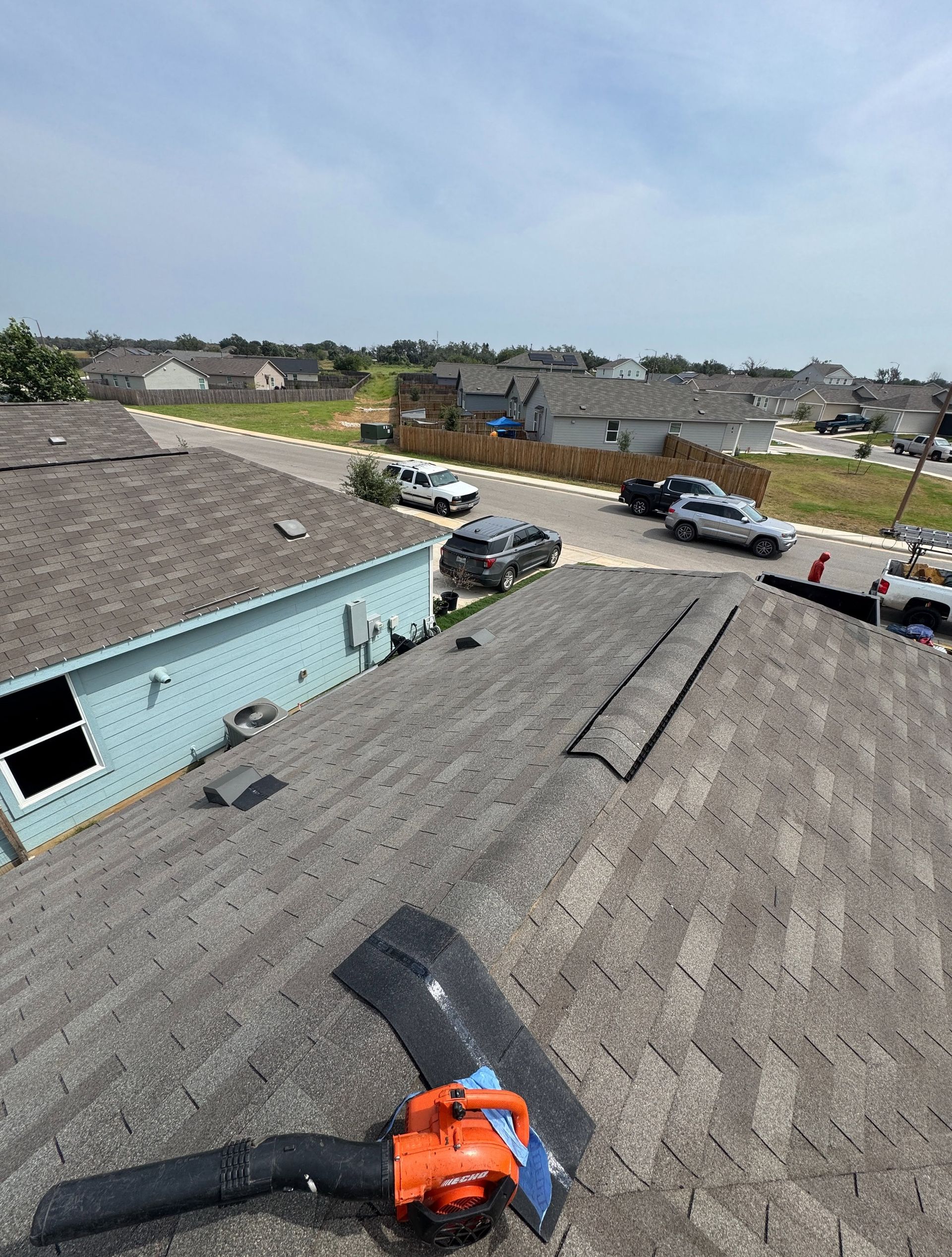 Roofer’s blower on gray shingle roof beside a light blue house, with suburban street and cars below.