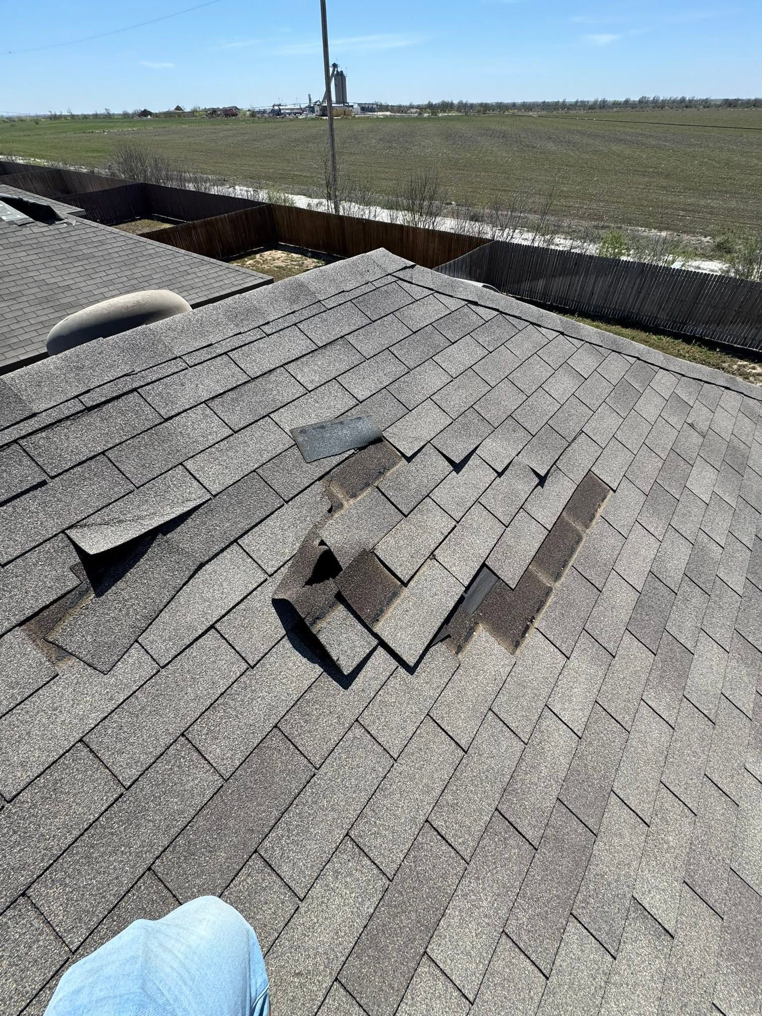 Damaged shingled roof with missing shingles and debris, viewed over farmland under a clear sky