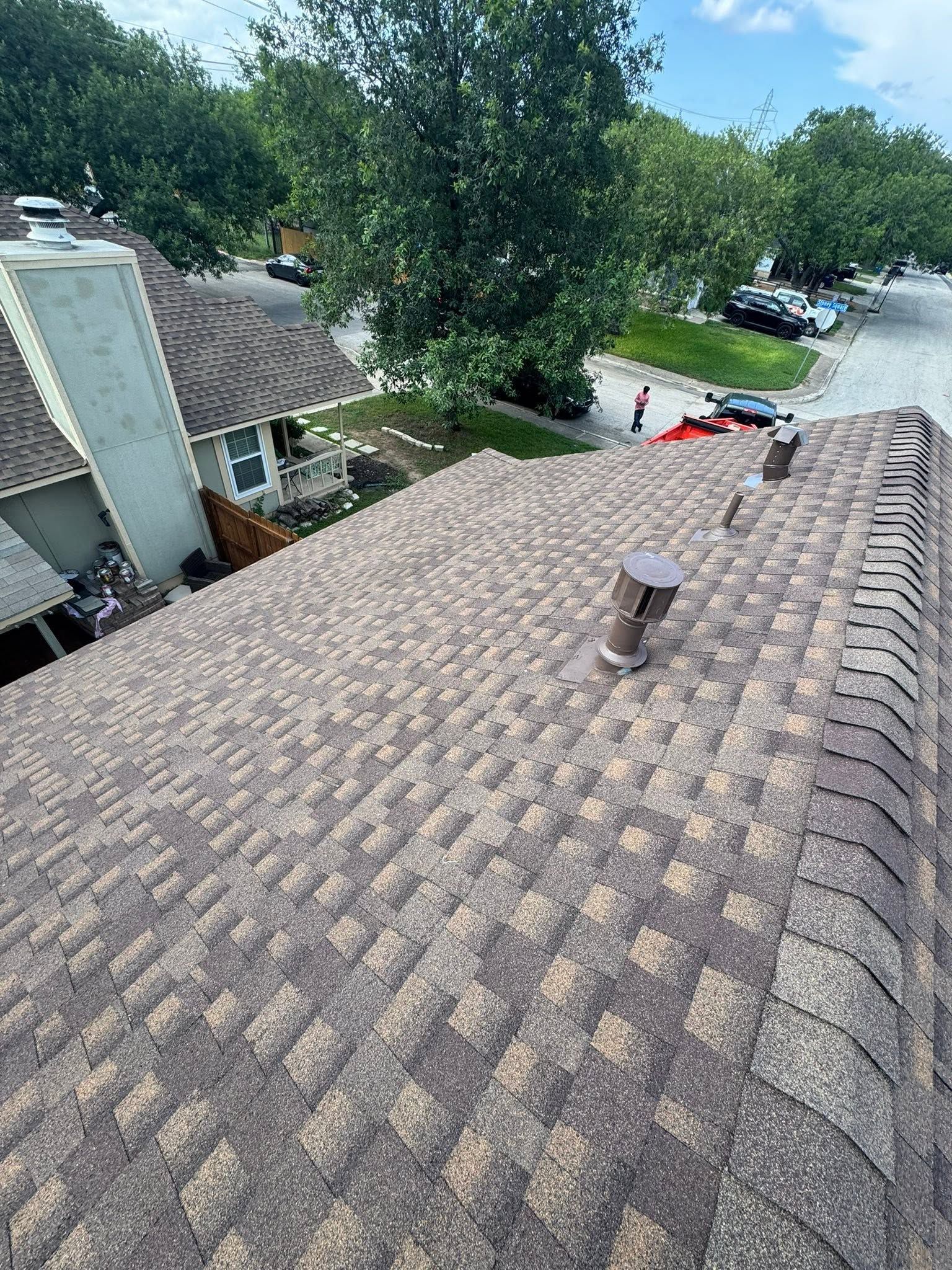 Rooftop with weathered shingles, vent pipe, and trees in the background near a suburban street.