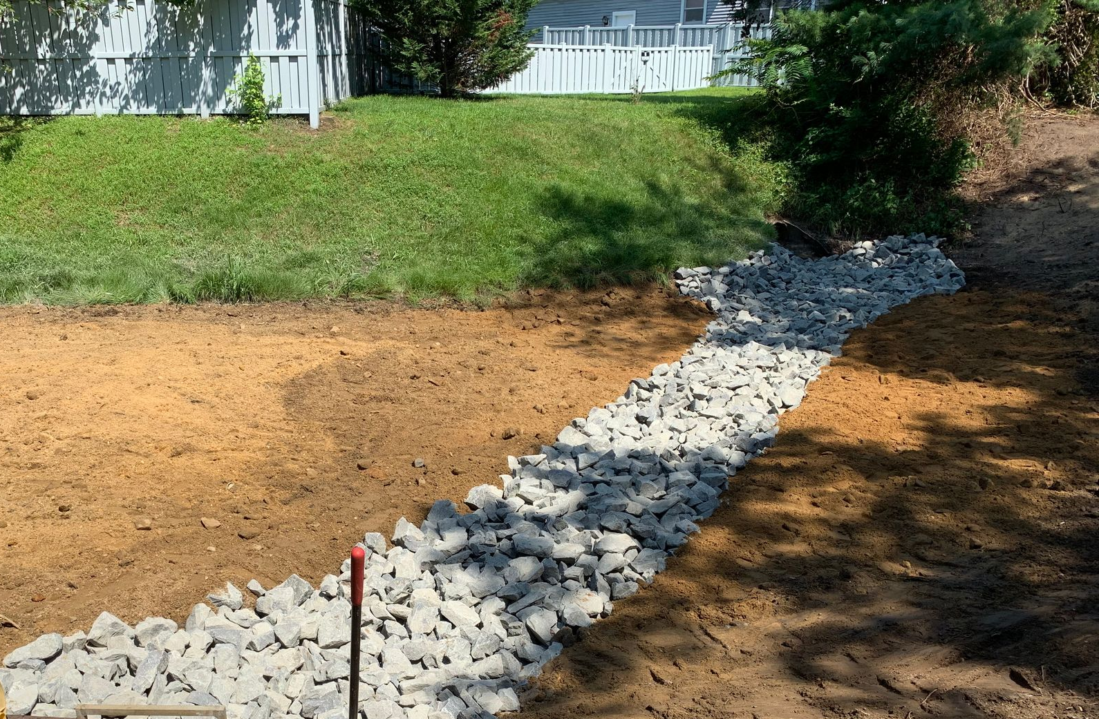 Backyard with newly graded soil and a gravel drainage trench beside a grassy lawn and white fence