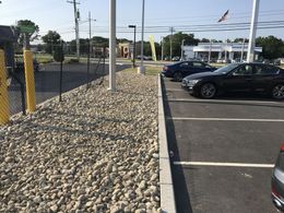 Rock-lined parking lot beside a chain-link fence, with parked cars and utility poles under a clear sky.