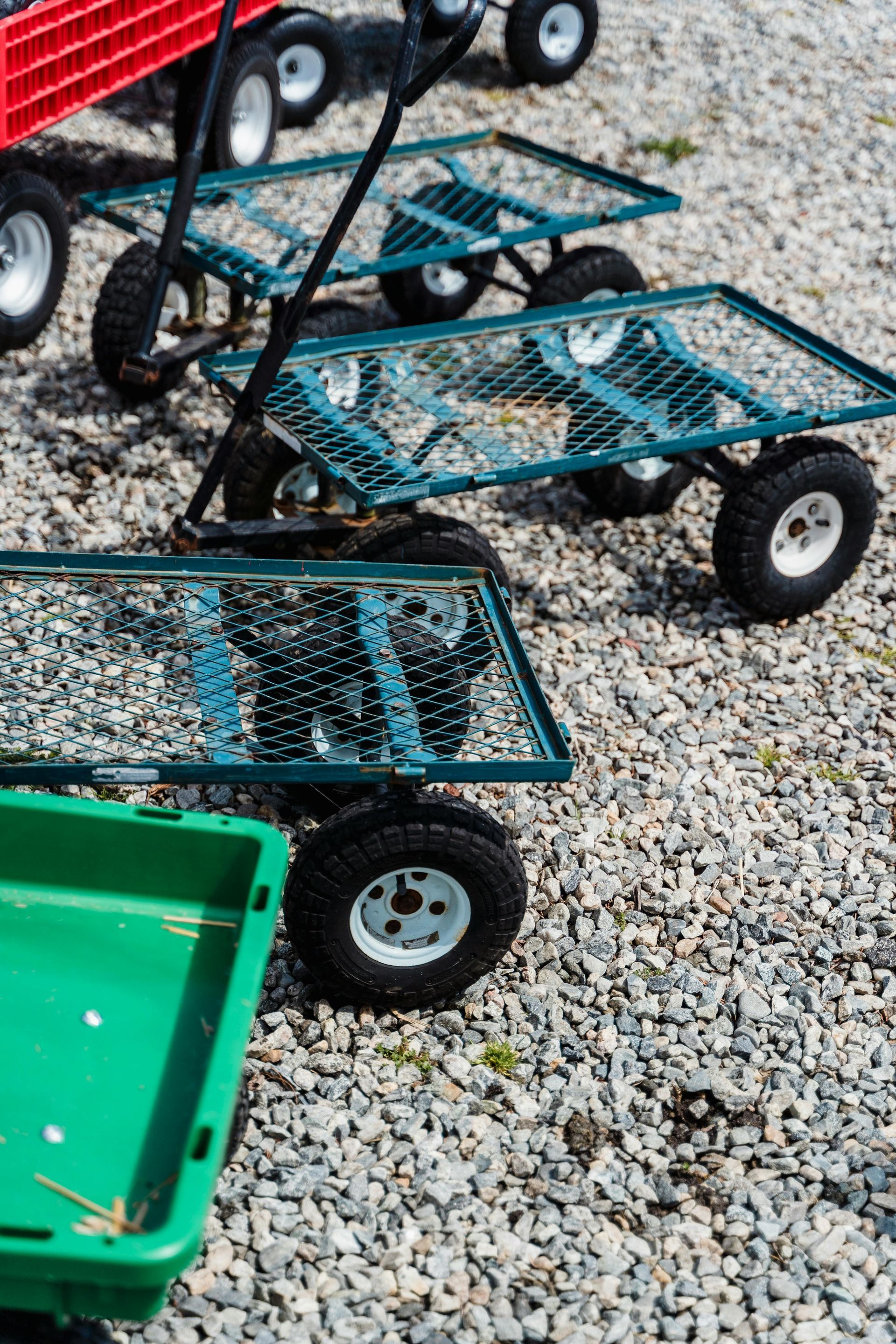 Stacked teal metal utility carts with black wheels on gravel, beside a green cart and red vehicle