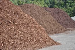 Piles of mulch in brown and purple hues stacked along a gravel roadside with trees in the background