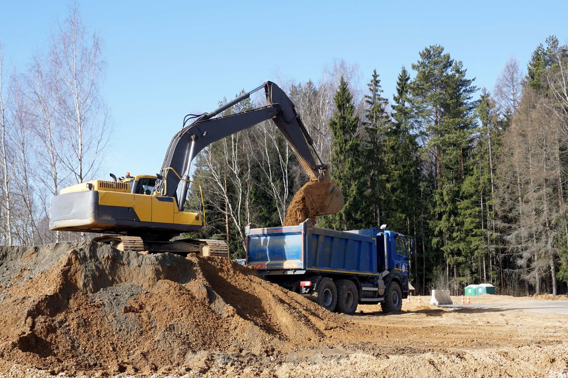 Excavator loading dirt into a blue dump truck at a construction site near trees