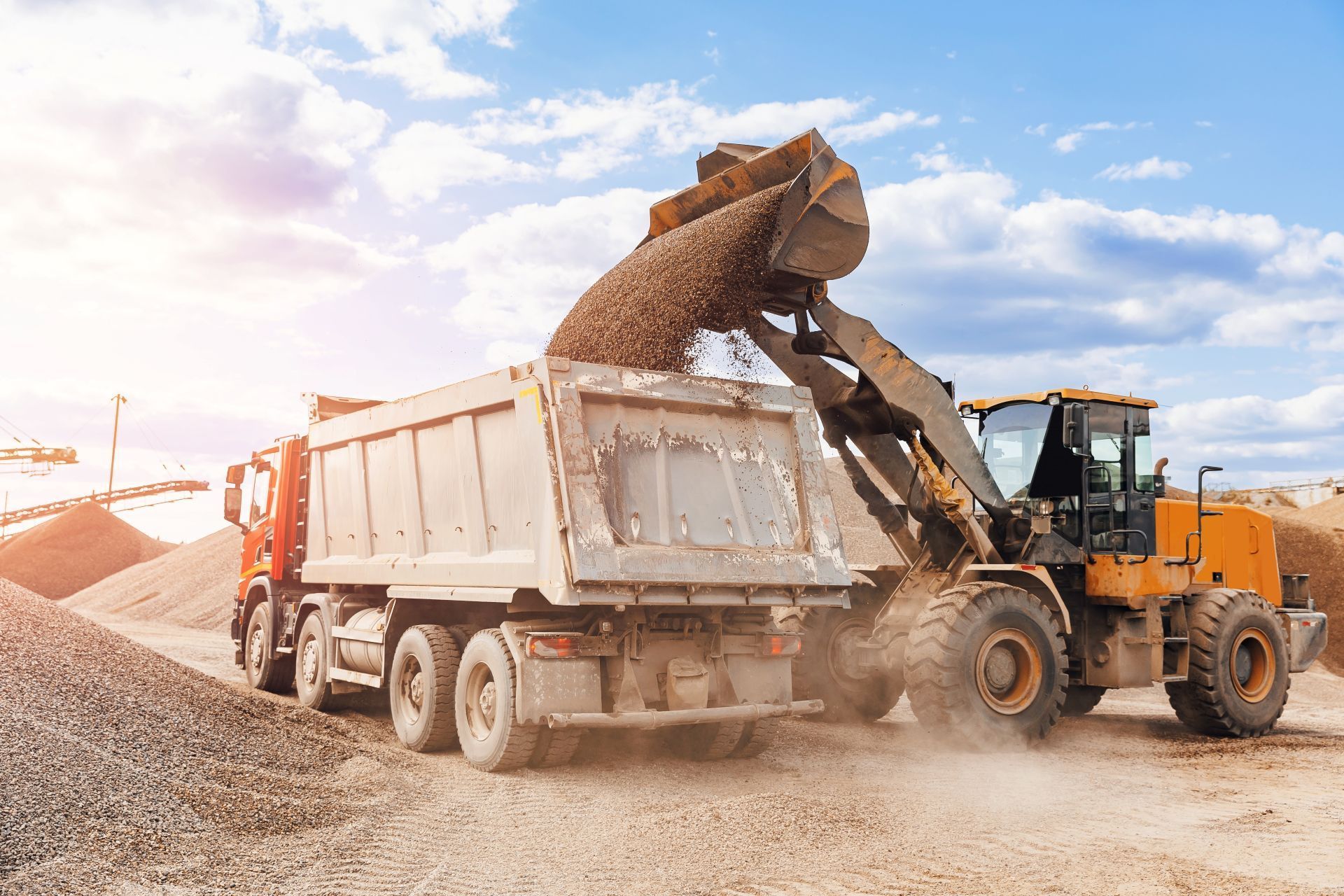 Yellow loader dumping gravel into a white dump truck at a construction site under a bright sky