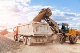 Yellow loader dumping gravel into a white dump truck at a construction site under a bright sky