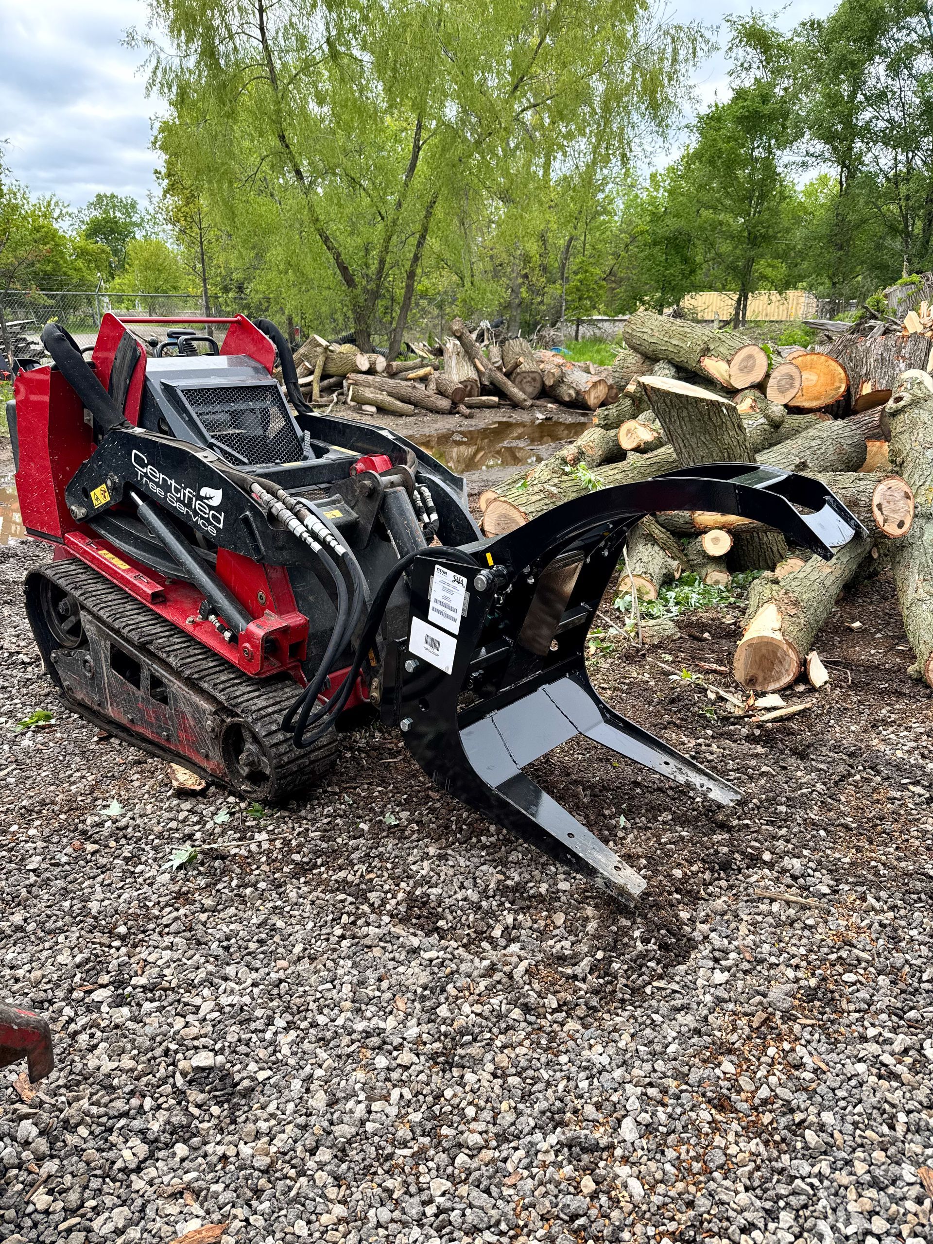 Red and black mini skid steer with grapple attachment, moving logs in an outdoor setting.