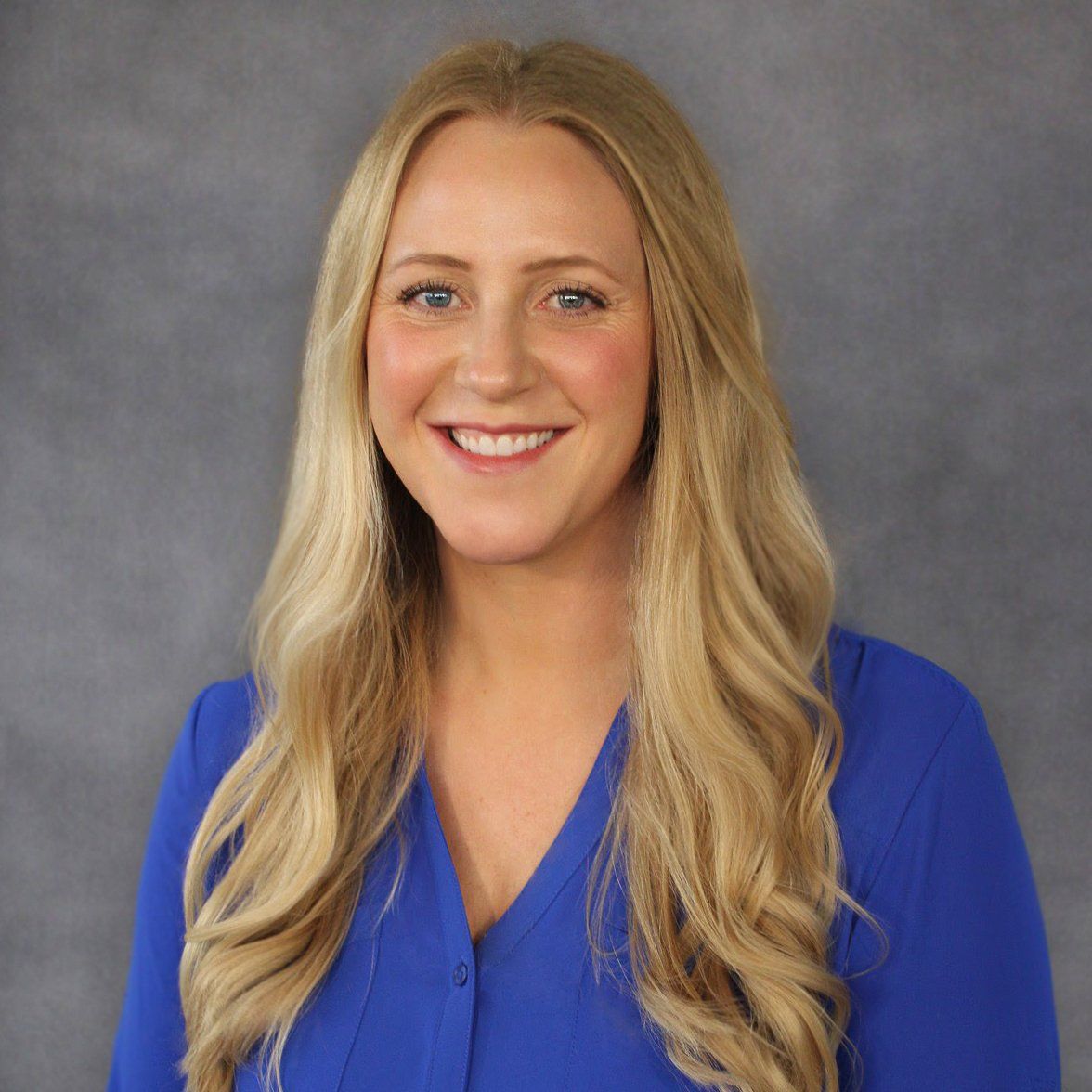 A woman with long blonde hair is wearing a blue shirt and smiling.