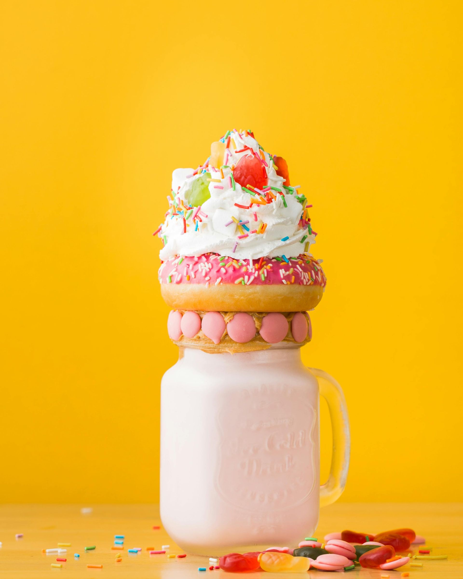 Pink milkshake in a jar, topped with a donut, whipped cream, sprinkles, and fruit, on a yellow background.