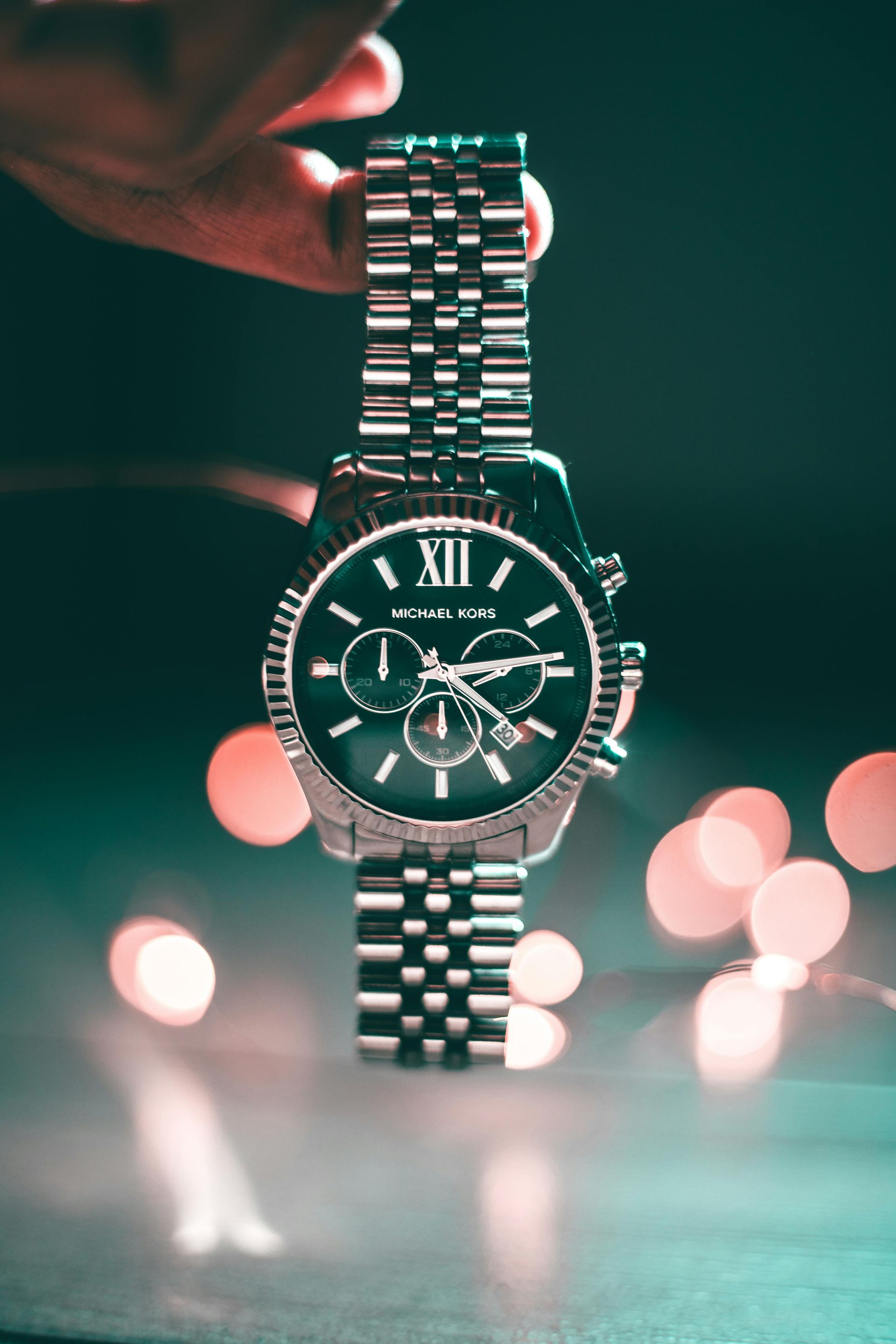 Silver Michael Kors watch held against a dark background with blurred lights.