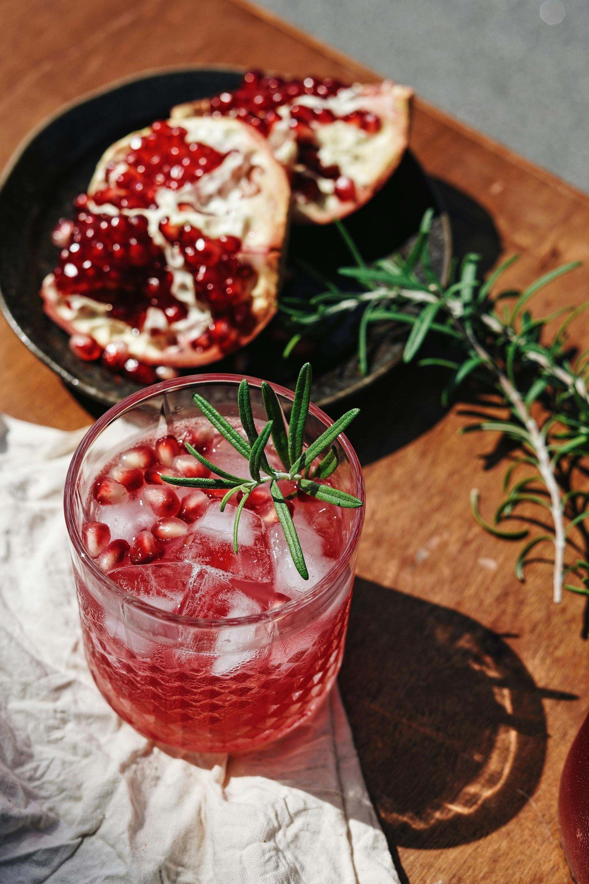 Cocktail with ice, pomegranate seeds, and rosemary sprig, beside pomegranate halves, on wood.